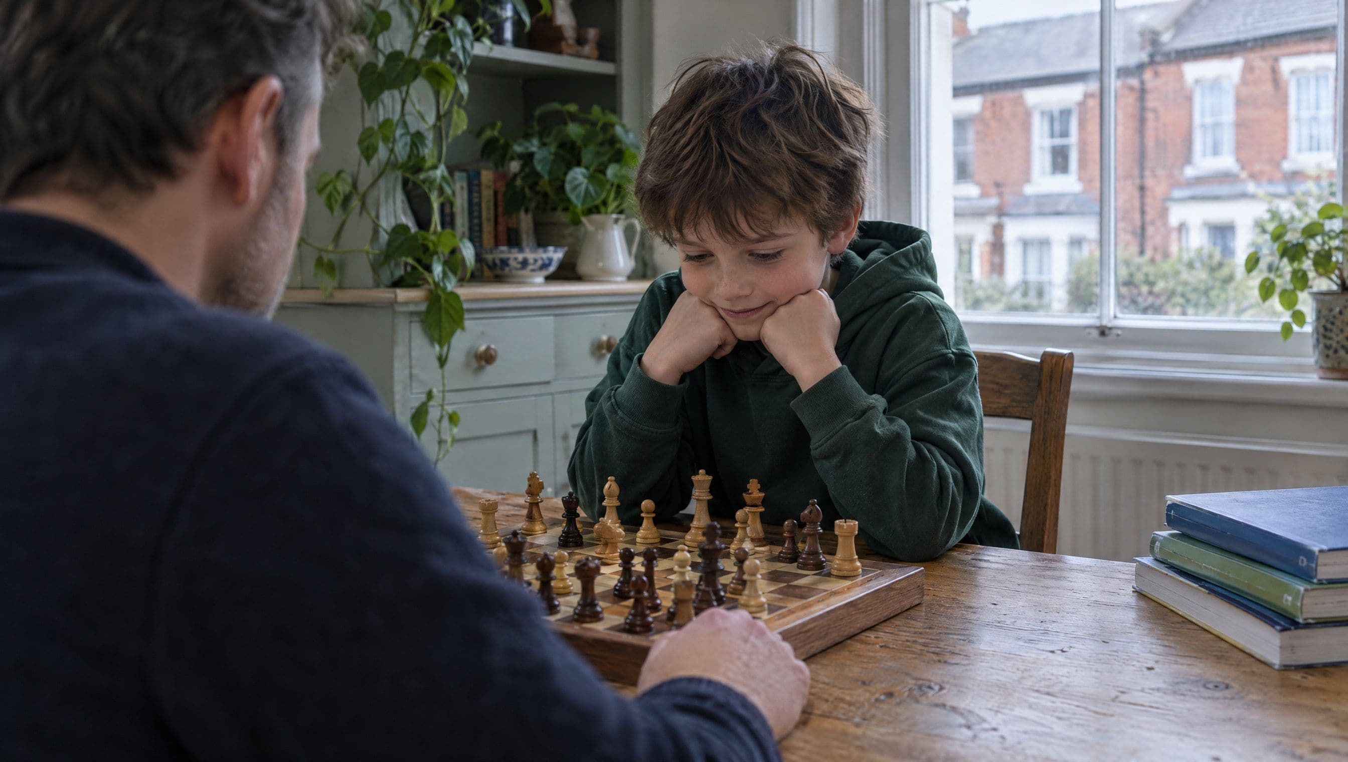 A boy leans over a chessboard with his chin on his fists, focused and half-smiling, as his father plays opposite.