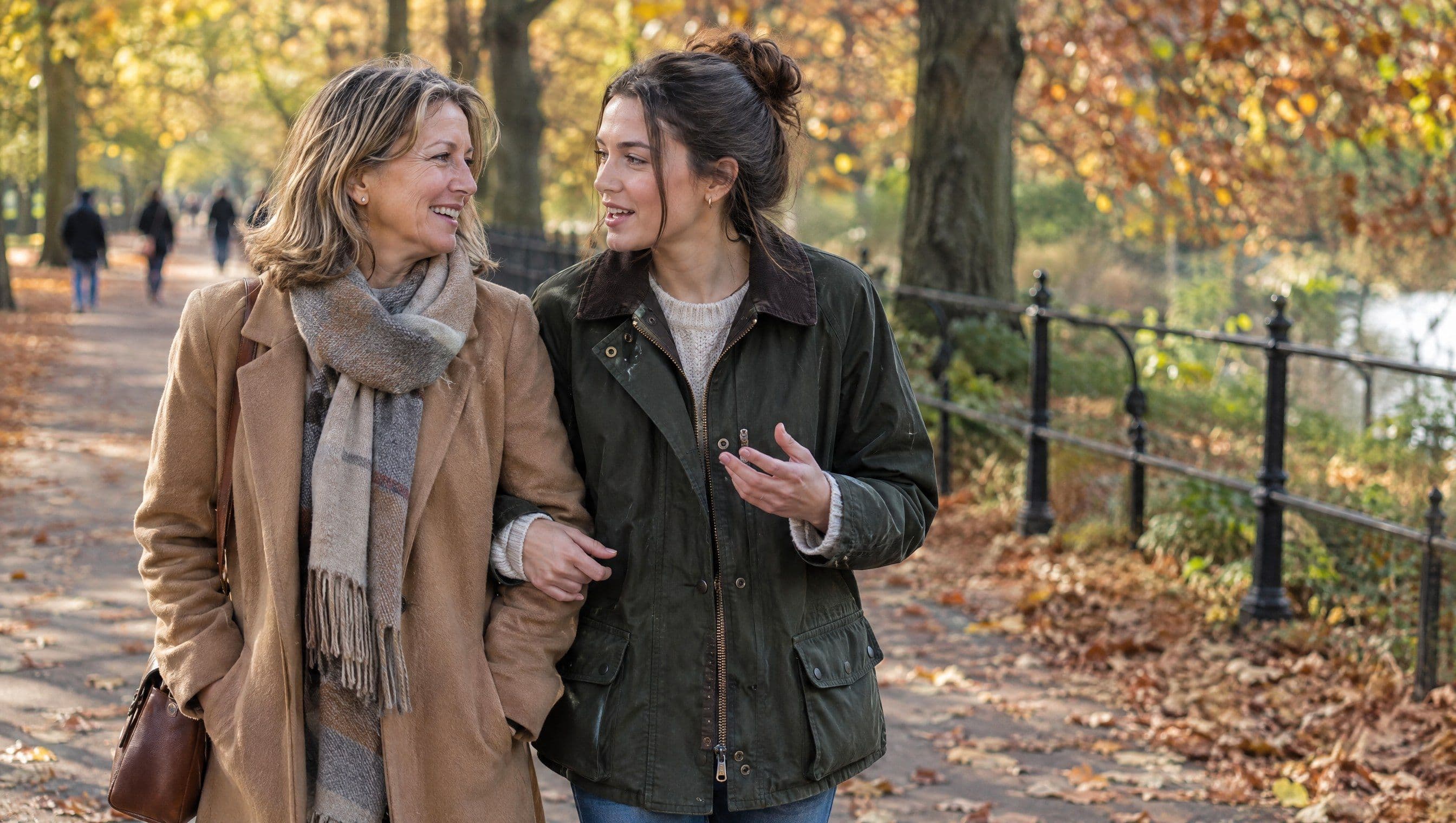 A woman in her forties and her adult daughter walk arm in arm along a park path in autumn, deep in conversation.