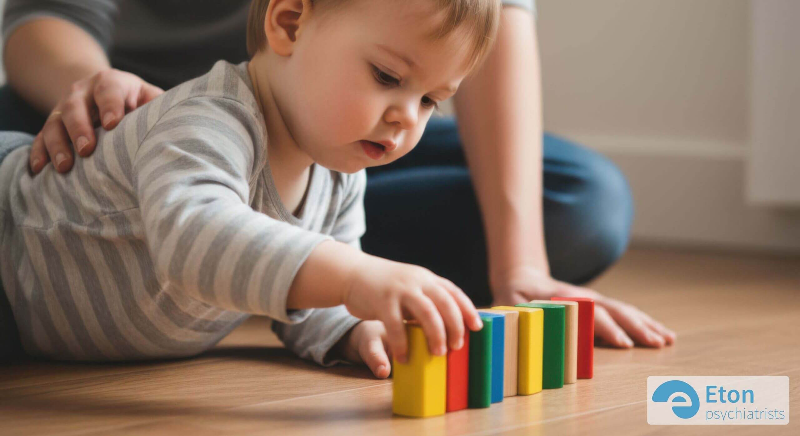 A parent's hands gently guide a toddler intently lining up colorful blocks, showing early focused play.